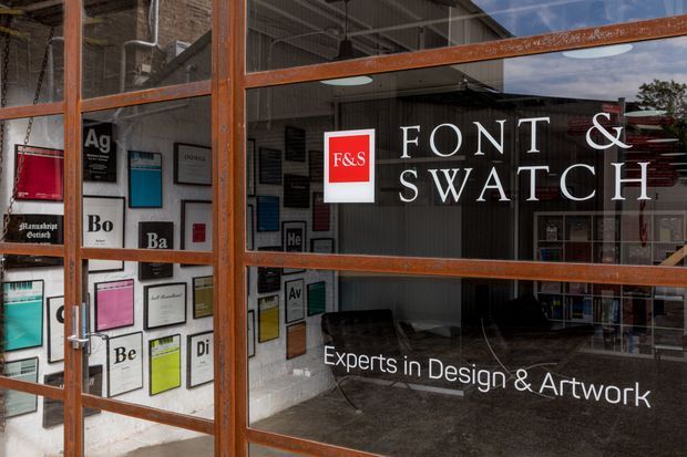 Exterior view through a rusted steel-framed glazed storefront into a graphic design and artwork studio. The facade displays large “Font & Swatch” lettering and an F&S logo, while framed typography and artwork samples are arranged on the interior walls.