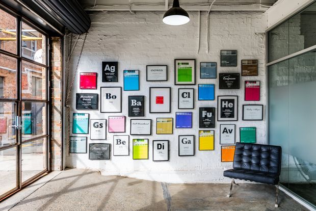 An industrial-style interior with exposed white-painted brick walls, polished concrete flooring, large black-framed factory windows, exposed ceiling services, and a black pendant light. The main wall is arranged as a gallery of framed graphic artworks inspired by periodic-table elements, with a tufted black leather lounge chair positioned in front. A glazed partition is visible on the right.