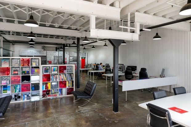 Open-plan industrial-style commercial office or studio with exposed white-painted roof trusses, beams, services and black structural columns. Corrugated metal wall cladding, polished concrete flooring, suspended black pendant lights, workstations, meeting tables, office chairs, and colorful shelving create a contemporary creative workplace atmosphere.