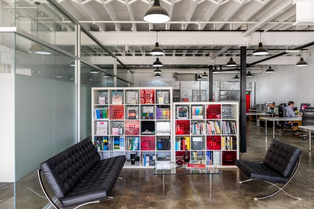 An open-plan contemporary office interior with exposed white structural beams and services, black pendant and track lighting, polished concrete flooring, glass-partitioned meeting or work rooms, and a central display shelving unit filled with books and magazines. Two black tufted lounge chairs create a reception or informal seating area in the foreground, while workstations are visible in the background.