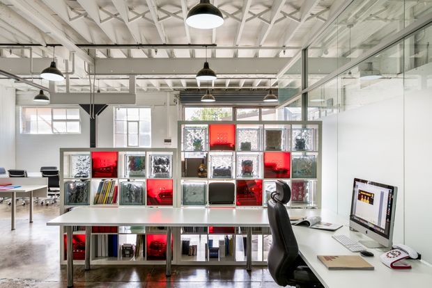 Bright open-plan creative studio or office with exposed white-painted roof trusses, polished concrete flooring, extensive internal glazing, suspended pendant lighting, and long white worktables. A striking modular shelving and display wall contains books, objects, and illuminated red compartments, with desktop computers and office chairs arranged around the workspace.