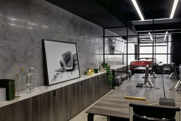 A sophisticated dark-toned commercial-style interior with a long timber-grain built-in cabinet and pale stone benchtop along a textured concrete-look wall. Black-framed glazed partitions divide the space, revealing desks and a red vehicle or product display beyond. Linear LED lighting runs through the black ceiling, while framed artwork, books, plants and sculptural objects add restrained decoration.