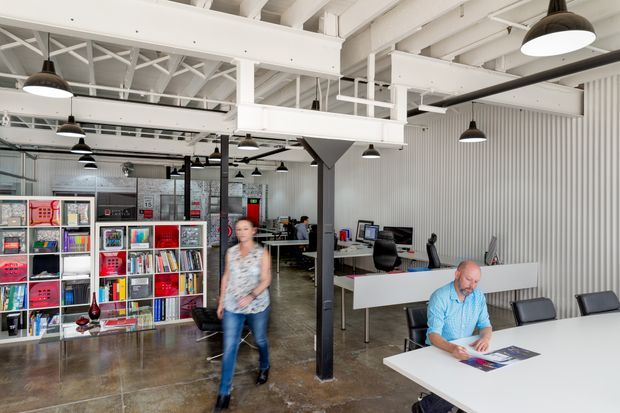 Open-plan commercial office and showroom with exposed white-painted steel roof trusses, beams, services, and mezzanine-like overhead structure. The space includes white corrugated wall cladding, polished concrete flooring, black pendant lights, long white worktables, office chairs, computers, and colourful shelving displaying printed materials or products. A person is walking through the foreground.
