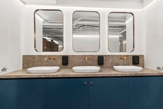 A contemporary bathroom vanity featuring three white vessel basins set on a brown stone benchtop, with three rounded-rectangle mirrors, warm brass wall-mounted taps, black fixtures, and dark navy-blue cabinetry. Exposed services are visible in the mirror reflections, suggesting a commercial or hospitality setting.