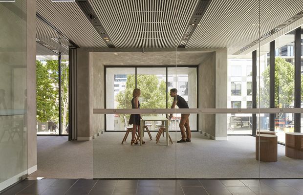 A contemporary commercial interior viewed through glazed entry doors, featuring an open lobby or reception area with polished dark floor tiles, grey carpet, exposed concrete-look columns and walls, extensive floor-to-ceiling glazing, and a linear slatted ceiling with recessed lighting. A small meeting table and stools sit centrally, with built-in timber-toned reception or display elements at the right. Urban buildings and trees are visible outside through the glazing.