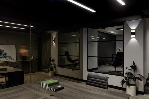 A dark, refined contemporary office interior with a large meeting table in the foreground, black leather office chairs, glass-partitioned private workspaces, and a lounge-style chair visible beyond the glazing. The space features a charcoal ceiling, linear recessed lighting, dark timber-look flooring, white structural walls, black-framed glass partitions, wall sconces, indoor plants, and subdued neutral finishes.