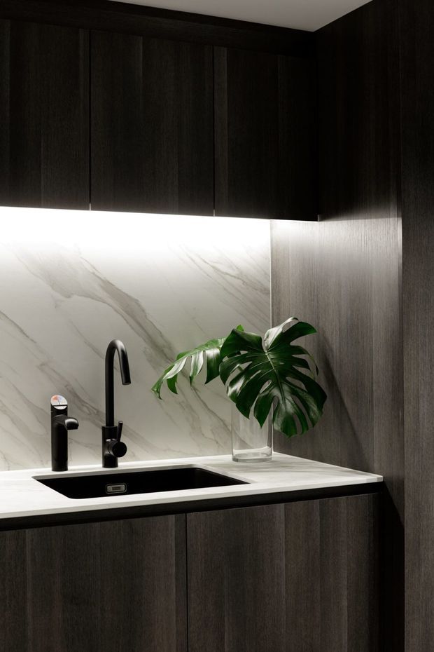 A compact, refined kitchen vignette featuring dark wood-grain cabinetry, a white veined stone backsplash and benchtop, an undermount black sink, and matte black tapware. Integrated under-cabinet lighting highlights the stone surface, while a clear vase with a tropical leaf adds a small natural accent.