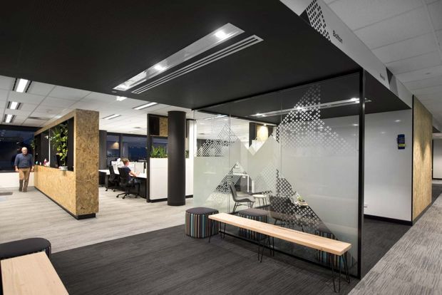 A contemporary commercial office interior with an open-plan workspace and a glass-enclosed meeting room. The office features patterned privacy film on the glazing, black acoustic ceiling panels with linear recessed lighting, grey carpet flooring, timber-faced partitions and columns, and a casual waiting area with a long timber bench and small round stool. Workstations and planted partitions are visible in the background.