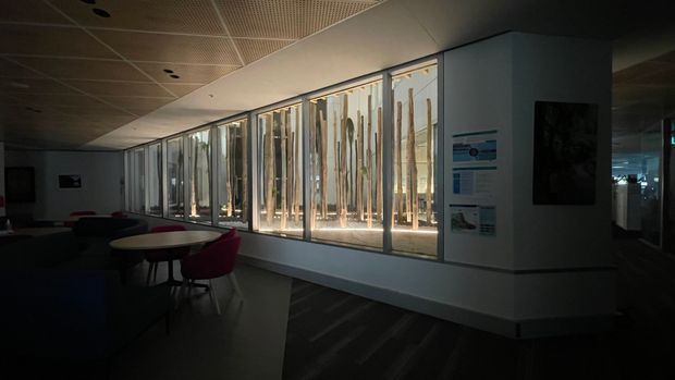 A dimly lit contemporary commercial interior, likely a lounge or waiting area, with low modular seating and small round tables arranged beside a long glazed wall. Vertical timber-like screening elements are visible beyond the glazing, while a perforated suspended ceiling, dark flooring, framed artwork, and wall-mounted informational displays define the space.