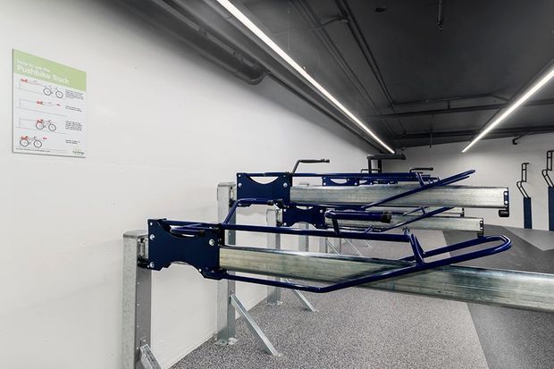 Interior view of a dedicated bicycle storage area with multiple wall-mounted and freestanding metal bike racks arranged along a white wall. The space has an exposed dark ceiling with visible services, linear strip lighting, a speckled grey floor, and a bicycle parking instruction sign.