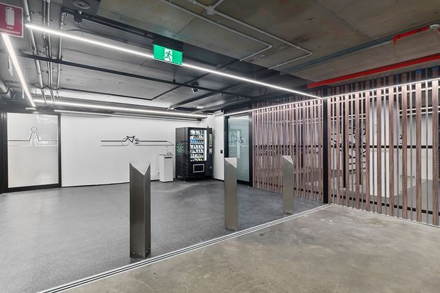 A modern commercial building lobby or access area with exposed concrete ceilings, visible services, linear LED lighting, polished concrete flooring, stainless-steel access bollards, and a vertical timber slat screen. A vending machine, bicycle storage signage, and emergency exit signage are visible, suggesting a shared office or apartment building circulation space.