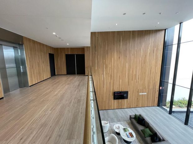 A contemporary multi-level lobby or circulation space with extensive vertical timber wall panelling, matching timber-look flooring, recessed ceiling lights, black-framed glazing, elevators, and a glass balustrade overlooking a seating area below. The lower level contains lounge seating and a small round table.