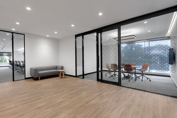 Contemporary open office interior with a spacious timber-look floor, white walls and ceiling, recessed downlights, and black-framed glazed partitions. A lounge area with a grey sofa and side table occupies the foreground, while a glass-enclosed meeting room with a conference table and rolling chairs is visible behind patterned privacy glazing.