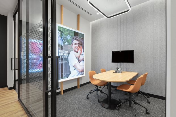 A compact contemporary meeting room or consultation room with a round light-wood table, four orange upholstered office chairs, wall-mounted display, desk phone, grey acoustic wall panels, dark carpet, and a black-framed glass partition. A large poster and timber accents add visual interest to the otherwise neutral commercial interior.