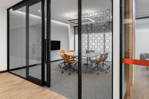 A contemporary commercial office interior viewed through black-framed glazed partitions. The meeting room contains a conference table with orange office chairs, grey carpet, patterned privacy film on the glass, linear ceiling lighting, and a wall-mounted screen. A red horizontal privacy stripe and glimpses of adjacent workspace are visible.