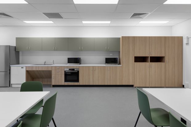 A contemporary office breakout kitchen and dining area with a long run of timber-look cabinetry, integrated appliances, a stainless-steel sink, grey overhead cupboards, and a tall timber-look storage unit with an open display niche. White tables and green chairs occupy the foreground beneath a suspended acoustic tile ceiling with recessed fluorescent lighting.