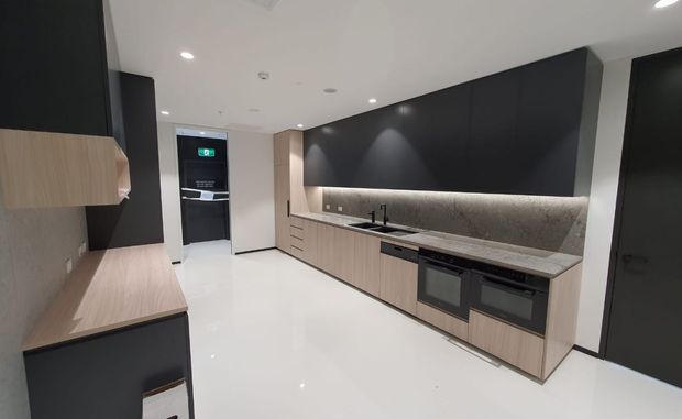 Contemporary galley-style kitchen with extensive matte black cabinetry, light timber-look lower cabinets, a long stone benchtop and integrated cooktop and double ovens. A textured grey stone splashback is illuminated by concealed strip lighting, while glossy white flooring and recessed ceiling lights create a clean, high-contrast finish.