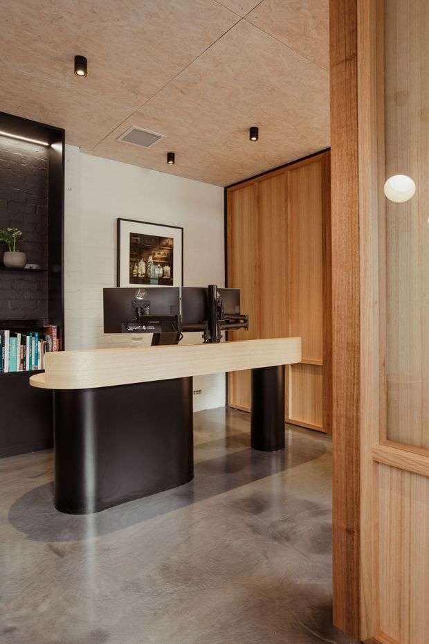A refined modern reception or administrative workspace with a curved pale timber desk supported by black cylindrical bases, dual monitors, and integrated shelving. The room features warm vertical timber wall panelling and a matching timber-framed glazed partition, a white painted masonry wall, black shelving with books and decor, a polished grey concrete floor, and a pale timber panel ceiling with recessed spotlights and ventilation. The composition is clean, warm, and highly designed.