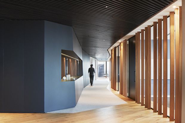 A refined contemporary interior corridor or lobby with a long linear perspective. The space features a dark ribbed ceiling, dark blue-grey sculptural wall and built-in display niche on the left, pale carpet through the circulation zone, warm timber flooring in the foreground, and repeated vertical timber screens with glazed openings on the right. Strong concealed and linear lighting creates a polished, high-contrast commercial or hospitality atmosphere.