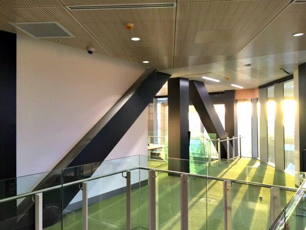 Interior view of a contemporary commercial circulation area or atrium with glass balustrades, green carpet, exposed black structural braces, extensive glazing, recessed lighting, and a perforated acoustic ceiling. The space appears to be an elevated hallway or landing overlooking a lower level.