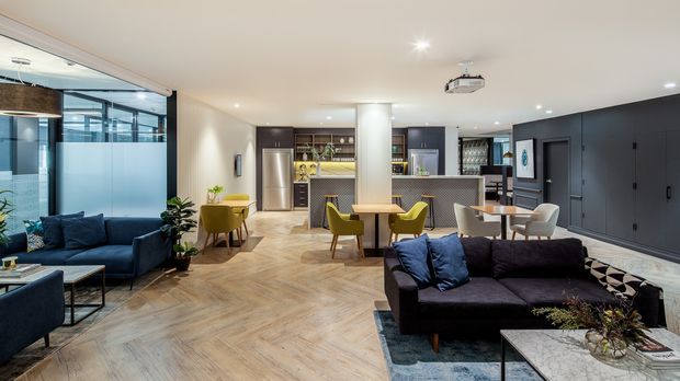 Open-plan contemporary living, dining and kitchen space with a dark built-in bar and cabinetry wall, patterned feature flooring, multiple seating areas, and a ceiling-mounted projector. Glazed partitions and a dark blue sectional sofa frame the bright, sophisticated interior.