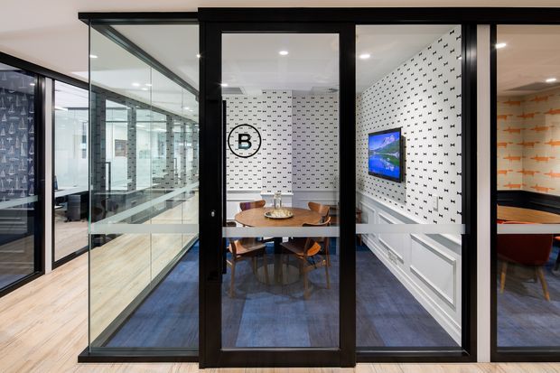 Contemporary office interior viewed through black-framed glazed partitions, with a small meeting room centered in the frame. The meeting room has a round timber table, curved timber chairs, blue carpet, patterned white walls featuring a circular logo, and a wall-mounted screen. Adjacent glazed rooms and a partial recreation or breakout area are visible to either side.