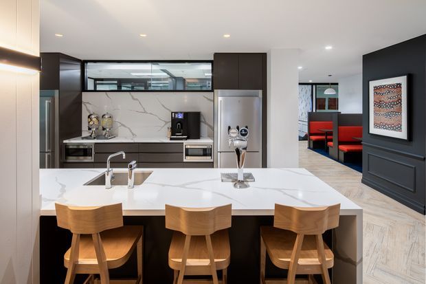 Contemporary open-plan kitchen with a large white veined stone island and three timber bar stools. Dark cabinetry contrasts with the pale benchtops and herringbone timber flooring. A black-panelled dining or lounge area with red upholstered seating is visible beyond the kitchen, along with integrated appliances, pendant lighting, and framed artwork.