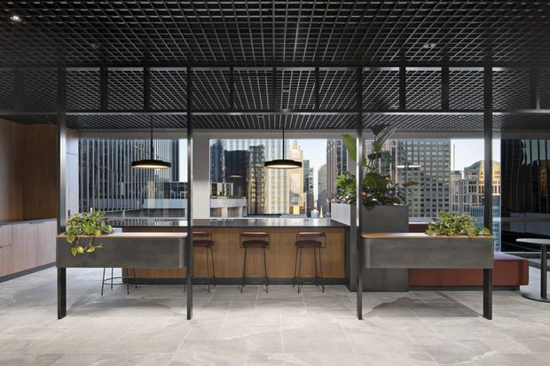A sophisticated open-plan commercial interior with a compact bar or kitchenette beneath a black open-grid ceiling. The space features timber and dark metal cabinetry, a stone-look floor, pendant lighting, planter boxes, and extensive glazing overlooking a dense city skyline. High stools line the central counter, with additional seating visible at the right.