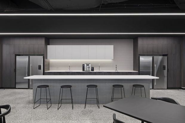 A contemporary commercial-style kitchen and dining space with a long central island, five black wire-frame bar stools, integrated appliances, dark timber-toned cabinetry, a pale tiled splashback, and a large dark dining table in the foreground. Linear strip lighting and an exposed dark ceiling create a refined industrial atmosphere.