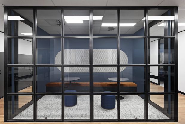 A contemporary commercial meeting room viewed through a black-framed glazed partition. The room contains a long warm timber meeting table, blue upholstered stools, a large light rug, and a patterned feature wall with rounded graphic forms. Reflections and views through the glazing reveal an open-plan office environment with a suspended acoustic-tile ceiling and recessed lighting.