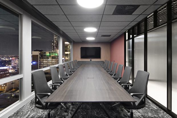 A contemporary corporate boardroom with a long rectangular conference table, rows of grey upholstered chairs, a wall-mounted display, floor-to-ceiling city-view glazing, and black-framed glass partitions. The room has a suspended grid ceiling with recessed lighting and a dark patterned carpet, creating a refined urban office setting.