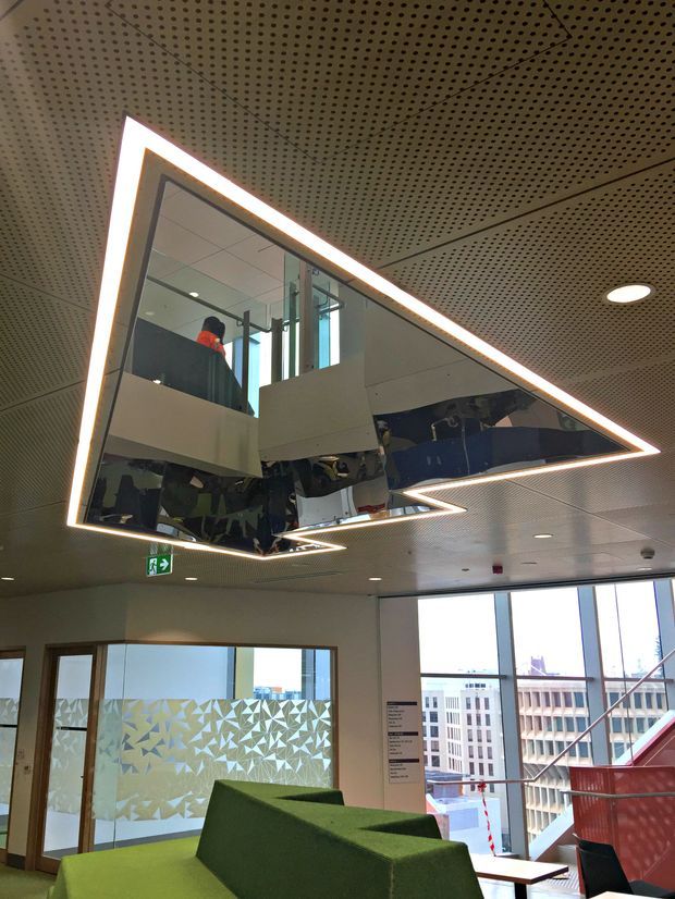 A contemporary commercial interior lobby or waiting area with bright green upholstered seating, extensive floor-to-ceiling windows, and a large angular mirrored ceiling feature outlined with integrated LED lighting. The ceiling has a perforated acoustic panel finish, while glazed partitions and a red stair or balustrade are visible in the background. Reflections in the mirrored ceiling show the surrounding interior and upper-level circulation.