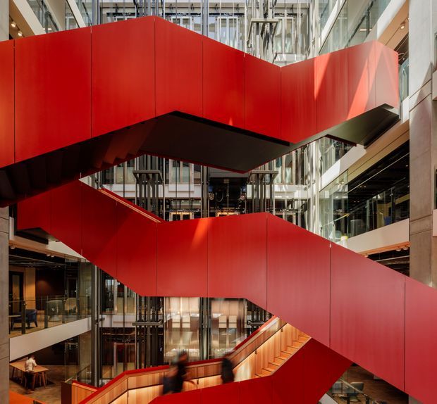 Interior view of a large contemporary commercial atrium dominated by a dramatic zigzagging red staircase or circulation bridge. The multi-level space has extensive floor-to-ceiling glazing, exposed services and structural elements, dark metal components, timber stair detailing, and abundant natural light.
