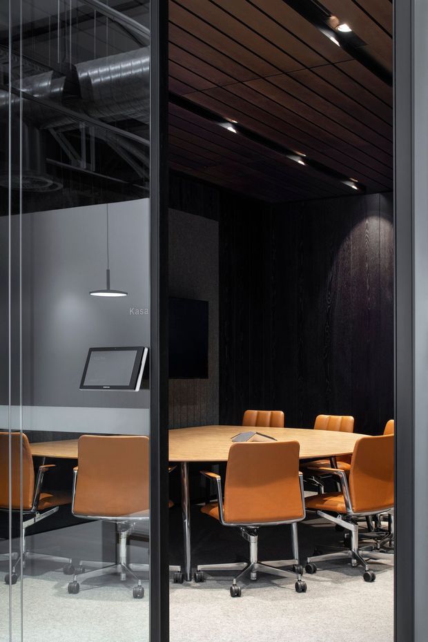 A dark, refined modern conference room viewed through a glazed partition. The room contains a large light-wood meeting table surrounded by tan leather or leather-look swivel chairs, with a wall-mounted display and a small table device. Black vertical timber wall panelling and a dark timber slatted ceiling with recessed spotlights create a dramatic interior. The glass partition also reveals exposed industrial ductwork and a pendant light in an adjacent area.