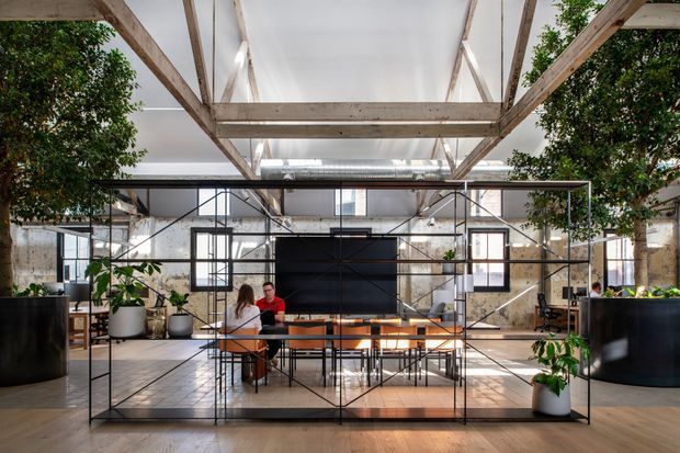 A bright, expansive open-plan interior with a large communal dining or meeting table enclosed by a black metal-framed glazed partition. The space features exposed timber roof trusses, industrial steel framing, high translucent ceiling panels, exposed ductwork, polished concrete floor areas, and aged masonry walls. Indoor trees and potted plants soften the industrial architecture, while several people occupy the central table.