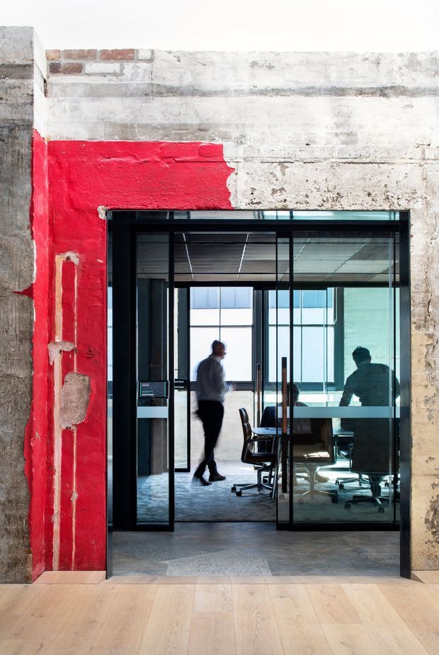 A contemporary commercial office interior viewed through a black-framed glass meeting-room entrance. The foreground has a light timber floor, while the surrounding walls retain a rough, weathered concrete-and-brick finish with a bold red painted section. Inside the glazed room are a meeting table, office chairs, large windows, and people working. The image strongly suggests an adaptive reuse or industrial-style workplace.