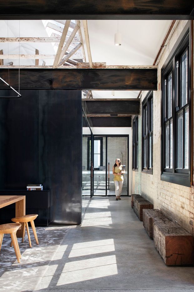 A bright industrial-style interior corridor or open-plan workspace with exposed timber roof framing, dark steel beams, black-framed windows and glazed partitions, painted white brick walls, and a polished concrete floor. Built-in or freestanding chunky timber bench seating lines the right wall, while a small timber desk and stool sit in the foreground. Strong sunlight creates geometric window shadows across the floor.