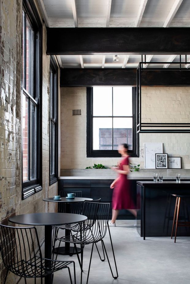 Industrial-style café or hospitality interior with exposed painted brick walls, large black-framed windows, dark steel structural beams, and a white exposed-beam ceiling. The space includes small round café tables with wire chairs in the foreground and a dark bar or service counter with stools at the rear. A blurred person is walking through the room.