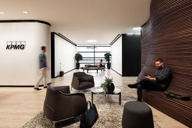 A contemporary commercial reception and waiting lounge branded KPMG. The open lobby features a curved illuminated reception wall, pale timber flooring, white and black partition walls, a large glazed window at the rear, dark leather lounge chairs, a low glass coffee table, and a dramatic curved wall clad in narrow dark timber slats with integrated bench seating. Neutral finishes and layered seating create a refined corporate atmosphere.