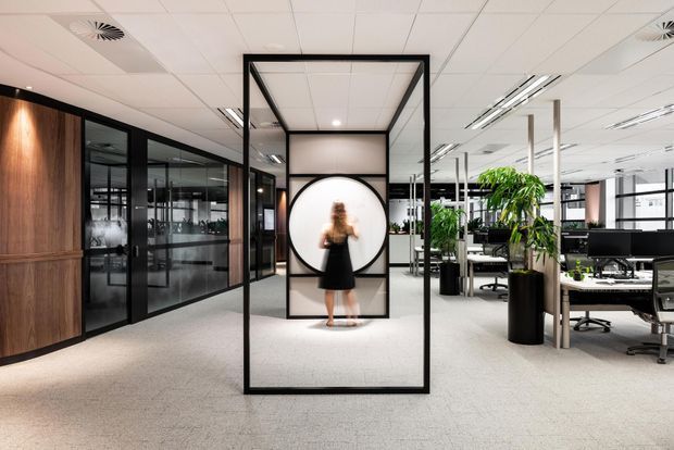 A contemporary open-plan commercial office interior with a reception or circulation area in the foreground. The space features black-framed glazed meeting rooms, warm timber wall panelling, carpeted flooring, suspended ceiling tiles with linear lighting, indoor plants, and rows of workstations. A sculptural black-framed glass partition with a circular feature opening forms the central architectural element.