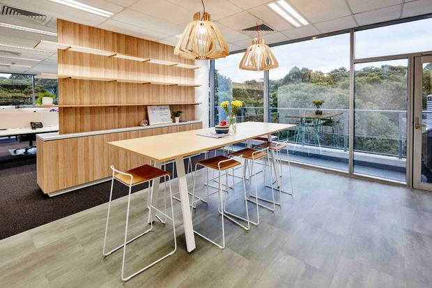 A bright contemporary office breakout and dining area with a long pale timber communal table, white-framed stools with brown seats, extensive floor-to-ceiling glazing, and a large built-in timber shelving and storage wall. Sculptural woven pendant lights hang above the table, while desks are partially visible to the left and greenery is visible beyond the windows.