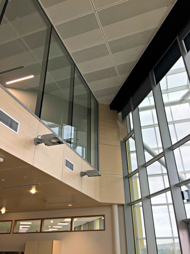 A tall contemporary commercial interior atrium or circulation space with a mezzanine level, exposed structural glazing, pale timber wall panels, suspended linear lighting, and a large floor-to-ceiling window wall. The space has a bright, open institutional or office-building character.
