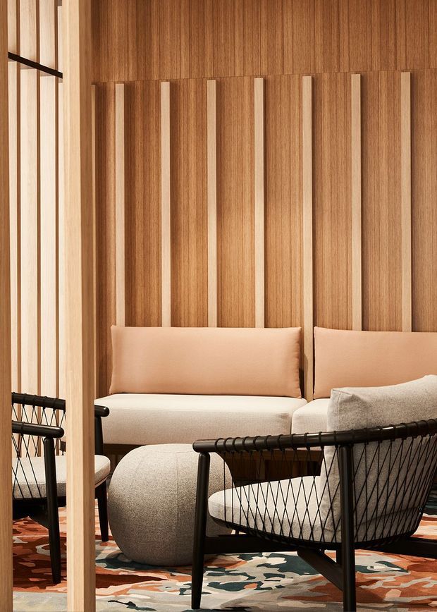 Warm contemporary lounge with a richly timber-panelled feature wall and evenly spaced vertical timber battens. The room contains a low upholstered sofa with peach cushions, rounded cream ottomans, black woven lounge chairs, and a patterned area rug in muted terracotta, cream, green and brown tones. Slim black framing and a timber screen or partition are visible at the left.
