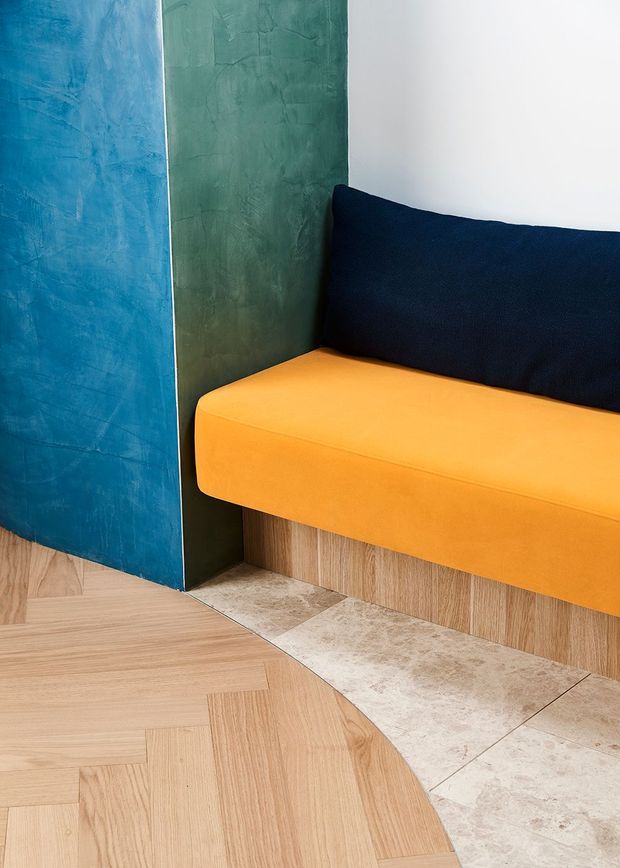 Close-up of a contemporary interior corner featuring a built-in orange upholstered bench with a dark navy back cushion, set against textured blue and green wall panels. Light timber herringbone flooring transitions to pale stone-look tiles, with timber-look fixed joinery beneath the seating.
