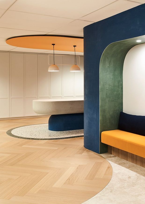 A refined contemporary interior featuring a curved reception or kitchen island, sculptural built-in navy and green arched joinery, orange upholstered seating and ceiling feature, pale timber herringbone flooring, and softly illuminated pendant lights. The space has rounded forms, integrated cabinetry, and a carefully coordinated blue, green, orange, cream, and light-wood palette.