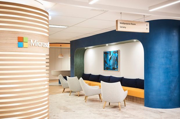 A bright contemporary commercial interior, likely an office reception or breakout lounge. A curved ribbed column with a Microsoft logo stands at left, while a deep blue curved feature wall frames a softly lit recessed seating nook with a long mustard-orange banquette, navy cushions, and several light upholstered lounge chairs. Directional signage for a multipurpose room and server area hangs from the ceiling. The space has a suspended ceiling, linear lighting, pale carpet, and strong blue, white, and warm yellow-orange accents.