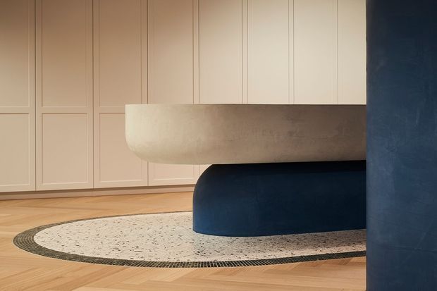A refined interior featuring a sculptural curved island or built-in bench with a pale textured upper form and deep navy rounded base. The room has warm herringbone timber flooring, a large oval terrazzo-like inset rug or floor feature with a dark mosaic border, and cream wall panelling with traditional rectangular mouldings. A matching navy curved column frames the right side of the image.