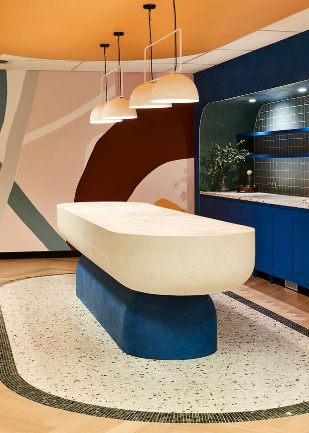 A bold, sculptural kitchen or bar interior featuring a large curved cream island with a deep blue rounded base, blue built-in cabinetry, and a dark blue tiled backsplash with open shelving. The room has a terrazzo floor with a contrasting mosaic border, colourful abstract wall artwork, an orange ceiling, and three oversized cream pendant lights. The overall design is highly expressive and contemporary.