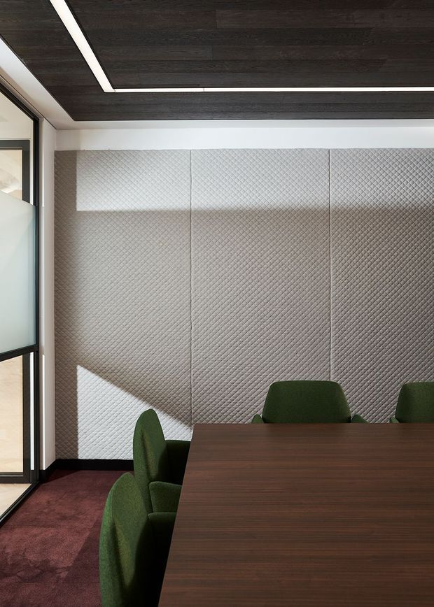 Contemporary meeting or dining room with a large dark timber table, green upholstered chairs, burgundy carpet, and a full-height quilted geometric wall panel. Glazing at the left brings in strong natural light, while dark timber ceiling panels with integrated linear lighting create a refined, high-contrast interior.