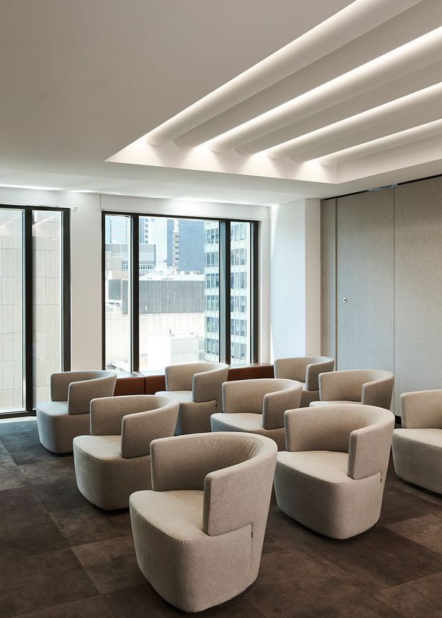 A bright, contemporary commercial lounge or waiting area furnished with numerous rounded upholstered armchairs arranged in small groupings. Floor-to-ceiling windows overlook a dense urban setting, while a large recessed ceiling feature with parallel rounded beams creates a distinctive architectural detail. The room uses a restrained palette of warm white, beige, taupe, and dark brown flooring, with a textured grey wall panel at right.